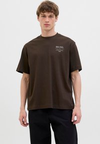 Dark brown cotton T-shirt with a relaxed fit, short sleeves, and a small white graphic print on the left chest area.