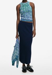 Top corto a maglia con motivo blu e verde, abbinato a una gonna lunga a coste blu scuro, e con un cardigan coordinato in mano. Scarpe nere con lacci.