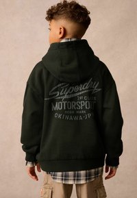 Tmavo zelená mikina s veľkým potlačeným logom na chrbte, ktorá obsahuje text "Superdry," "Motorsport" a "Okinawa, Japonsko." Nosená cez károvanú košeľu.