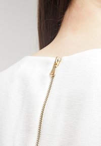 Arrière du cou d'une personne aux cheveux bruns visibles, portant un vêtement blanc texturé avec une fermeture éclair dorée partiellement fermée.