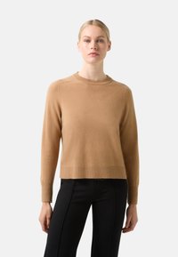 Pull en maille couleur camel avec des poignets et un ourlet côtelés, présentant un col rond et des coutures d'épaule discrètes. Associé à un pantalon noir.