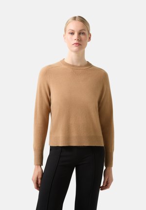 Pull en maille couleur camel avec des poignets et un ourlet côtelés, présentant un col rond et des coutures d'épaule discrètes. Associé à un pantalon noir.
