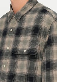 Camisa de cuadros en colores beige, negro y gris. Presenta un cuello, dos bolsillos en el pecho y botones blancos en una tela texturizada.