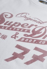 Hvid stof t-shirt med fed bordeaux tekst, inklusive "Original SuperDry Leather & Boot Co." med indviklede skrifttype designs og accenter.