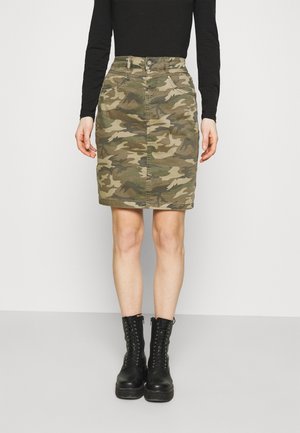 Kvinde iført en camouflage nederdel til knæet, sort langærmet top og sorte snørebåndsankelstøvler står foran en ensfarvet baggrund.