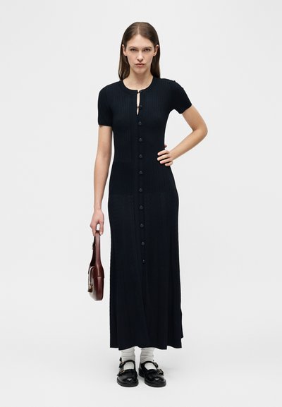 Γυναίκα που φοράει μαύρο μάξι φόρεμα με κοντά μανίκια και κουμπιά, λευκές κάλτσες, μαύρα παπούτσια Mary Jane, κρατώντας ένα καφέ τσάντα στο αριστερό της χέρι.