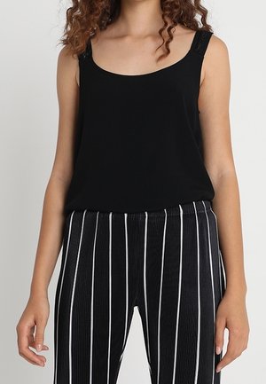 Nainen, jolla on sleeveless musta yläosa ja mustat housut, joissa on valkoisia pystysuoria raitoja, seisoo yksiväristä vaaleaa taustaa vasten.