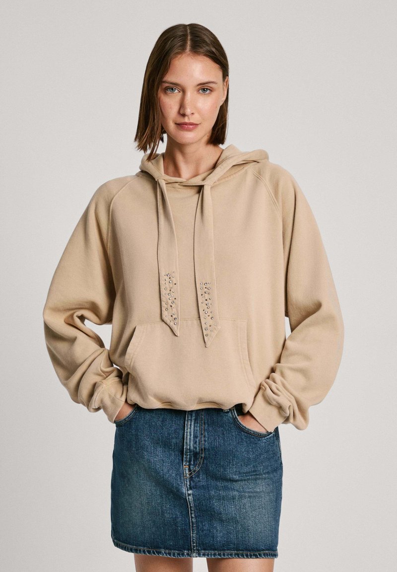 Sudadera con capucha beige con un bolsillo frontal y cordones decorativos. Llevada con una falda mini de mezclilla, mostrando un estilo casual.