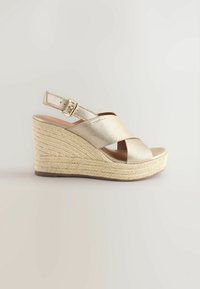 Sandale compensée en métal doré avec design entrecroisé, sangle ajustable et semelle en jute tissée ; texture lisse et semelle intérieure légèrement rembourrée.