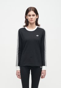 Mladá žena v čiernom tréningovom obleku Adidas s bielymi pruhmi na rukávoch a nohaviciach, stojaca pred jednoduchým bielym pozadím.