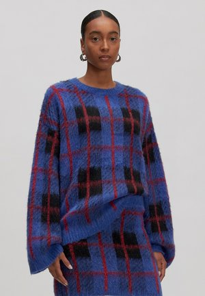 Een vrouw in een blauwe fuzzy trui en een rok met een rood en zwart ruitpatroon, staand voor een effen lichte achtergrond.