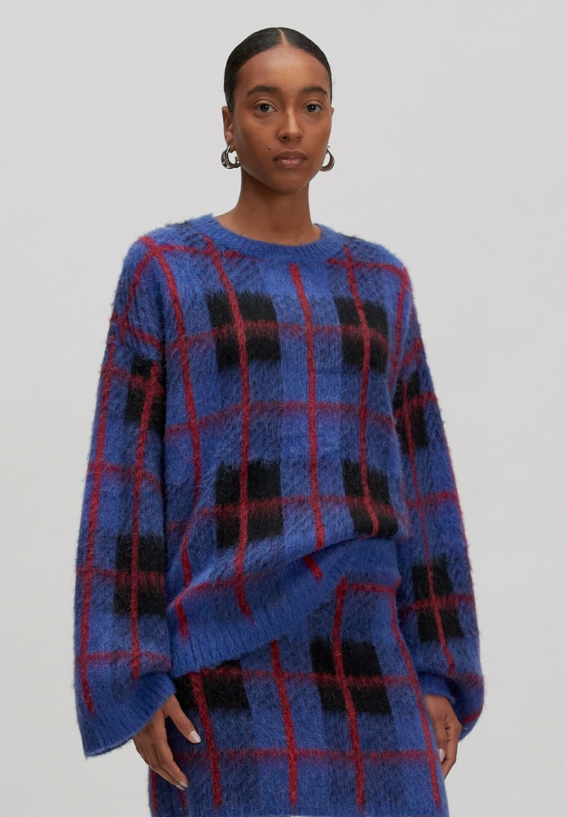 Frau in einem blauen flauschigen Pullover und einem Rock mit rot-schwarzen Karomustern, die vor einem schlichten hellen Hintergrund steht.