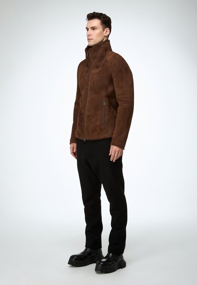 Braune Wildlederjacke mit hohem Kragen, Frontreißverschluss und zwei Seitentaschen, kombiniert mit schwarzer Hose und klobigen schwarzen Stiefeln.