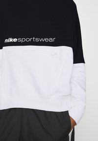 Nike-huppari, jossa on musta yläosa ja valkoinen alaosa, vetoketjullinen tasku ja rohkea logo edessä.