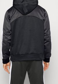 Sweat à capuche sportif noir avec une texture lisse, des bandes latérales blanches contrastantes et une capuche à cordon. Coupe slim avec poignets et ourlet côtelés.