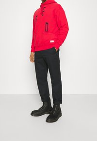 Sudadera con capucha roja y bolsillo frontal con letras negras, combinada con pantalones oscuros y botas negras de cordones. Conjunto casual sobre un fondo neutro.