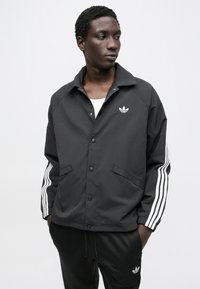 Črna športna jakna s ovratnikom, sponkami na pritrditev, stranskimi žepi in belimi tremi črtami na rokavih. Logotip Adidas na prsih.