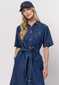 Femme aux longs cheveux blonds portant une robe en jean foncé boutonnée avec une ceinture nouée à la taille et une casquette en jean assortie, debout devant un fond uni.