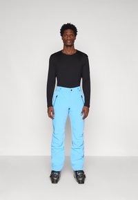 Juoda ilga rankovė palaidinė derinama su šviesiai mėlynomis slidinėjimo kelnėmis, turinčiomis šonines kišenes, vilkima su juodais slidinėjimo batais. Paprastas, prigludęs dizainas.