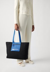 Svart tote-väska med blå detaljer, med en framficka med dragkedja och logo-text. Tillverkad av hållbart material; stor rektangulär form.