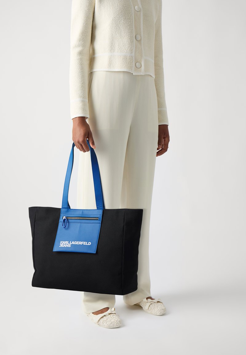 Svart tote-väska med blå detaljer, med en framficka med dragkedja och logo-text. Tillverkad av hållbart material; stor rektangulär form.