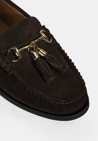 Donkerbruine suede instapper met een ronde neus, decoratieve gouden hardware en dubbele kwastaccenten. Beschikt over een gestikte rand en een zwarte zool.