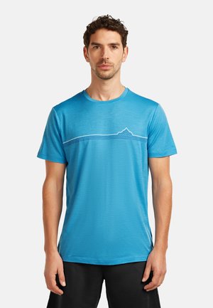 Mann mit kurzen lockigen Haaren, der ein blaues T-Shirt mit kurzen Ärmeln trägt, das eine weiße Linie und ein Bergdesign über der Brust hat, stehend vor weißem Hintergrund.