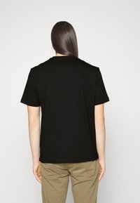 Persona con cabello largo y lacio, vistiendo una camiseta negra de manga corta lisa y pantalones caqui, de espaldas contra un fondo blanco.