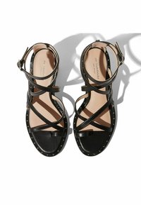 Crne kožne sandale s višestrukim ukriženim trakama, otvorenom nožnom dizajnom, ravnim potplatom i bočnim zakovicama. Imaju kopčanje na gležnju.