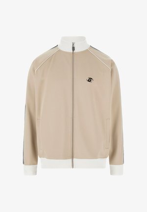 Beige trainingsjack met een hoge witte kraag, een ritssluiting aan de voorkant, zijzakken en zwarte accenten op de mouwen. Heeft een klein logo op de borst.