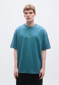 Teal υπερμεγέθη μπλουζάκι από μαλακό ύφασμα, με μικρό πράσινο λογότυπο "HUGO" στο στήθος. Κοντά μανίκια και σχέδιο με στρογγυλή λαιμόκοψη.