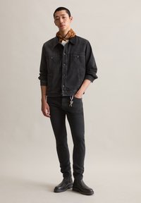 Zwarte denimjasje met voorzakken en knoopsluiting, gecombineerd met strakke zwarte jeans en zwarte enkelboots; heeft een bruine sjaal.