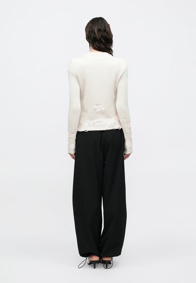 Pulover bej cu dungi, cu detalii uzate, asortat cu pantaloni largi negri și pantofi cu toc înalt, văzut din spate.