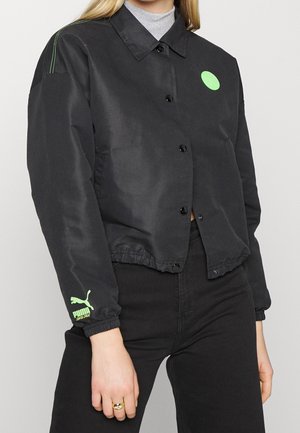 Kobieta ubrana w czarną kurtkę Puma z neonowo zielonym logo na rękawie i piersi, połączoną z czarnymi spodniami i szarym golfem pod spodem.