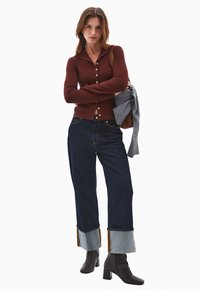 Brun langærmet skjorte med knapper, mørkeblå vidde jeans med denimmanchetter, og højhælede sorte støvler til knæene. Grå sweater draperet over armen.