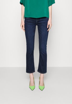 Mujer con vaqueros azul oscuro cortos, top verde y tacones altos de punta verde brillante, de pie sobre un fondo blanco.