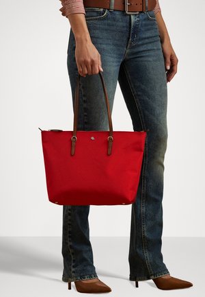 Red tote bag with brown leather handles, canvas material, featuring a logo detail on the front and a structured design, held by a person.