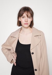 Trench coat beige com lapelas largas, botões pretos e um ajuste descontraído, usado sobre uma blusa preta, em frente a um fundo neutro.