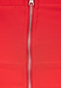 Gros plan sur une fermeture éclair en métal argenté sur un tissu rouge vif avec des coutures visibles le long des bords de la fermeture.