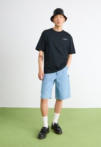 Black cotton t-shirt with a small logo, light blue denim shorts, white socks, black shoes with tassels, standing on green flooring.