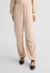 Pantaloni larghi beige chiaro con vita regolabile, realizzati in un tessuto texturizzato e presentano polsini con zip, abbinati a scarpe nere con punta affilata.
