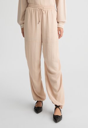 Pantaloni larghi beige chiaro con vita regolabile, realizzati in un tessuto texturizzato e presentano polsini con zip, abbinati a scarpe nere con punta affilata.