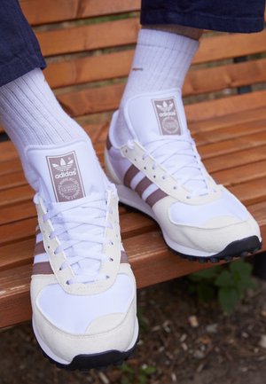 Chaussures de sport blanches en cuir texturé et mesh, avec trois bandes brunes, des lacets blancs et une étiquette logo sur la languette.