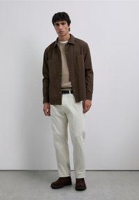 Bruin overhemd met knopen over een beige trui, gecombineerd met gebroken witte broek en bruine laarzen. Het overhemd heeft twee voorzakken.