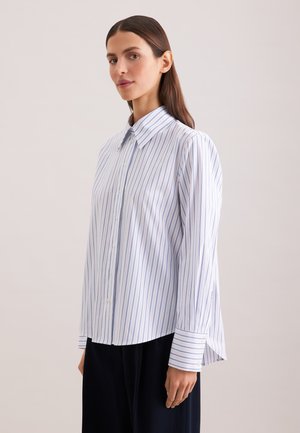 Femme aux cheveux bruns et raides porte une chemise blanche à fines rayures verticales bleues et un pantalon noir, debout devant un fond uni.