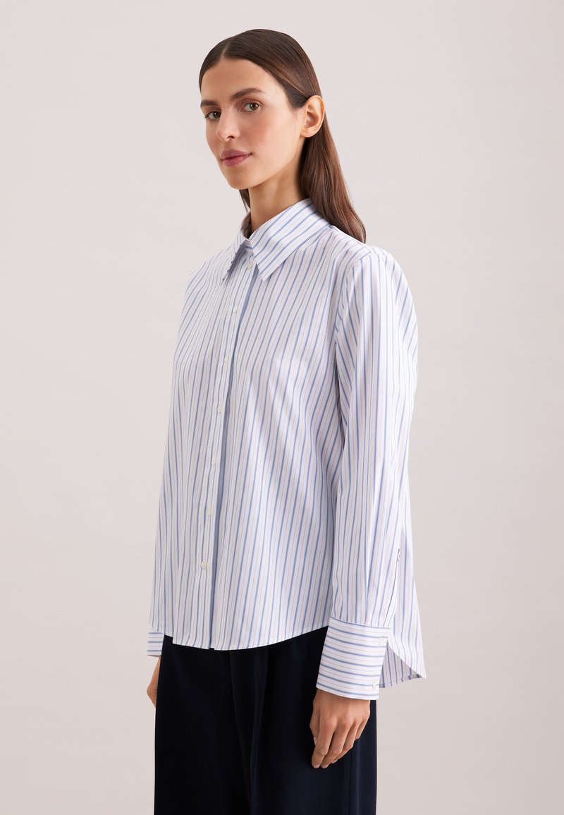 Femme aux cheveux bruns et raides porte une chemise blanche à fines rayures verticales bleues et un pantalon noir, debout devant un fond uni.