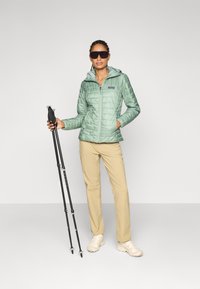 Lichtgroene gewatteerde jas met een capuchon, gecombineerd met beige broek en crèmewitte schoenen. Houdt wandelpalen met zwarte handvatten vast. Zonnebril op.
