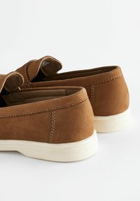 Brązowe zamszowe buty o gładkiej powierzchni, zaokrąglonych krawędziach i białych gumowych podeszwach. Podwójny design wsuwany z przeszyciami.
