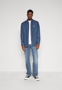 Chemise en denim bleu moyen avec fermeture à boutons et poche poitrine simple, associée à un jean bleu clair et des baskets noir et blanc.