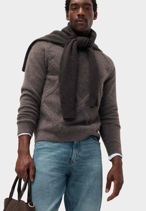 Mężczyzna w brązowym swetrze z szalem zawiązanym na szyi, w jasnoniebieskich dżinsach, trzymający brązową torbę.